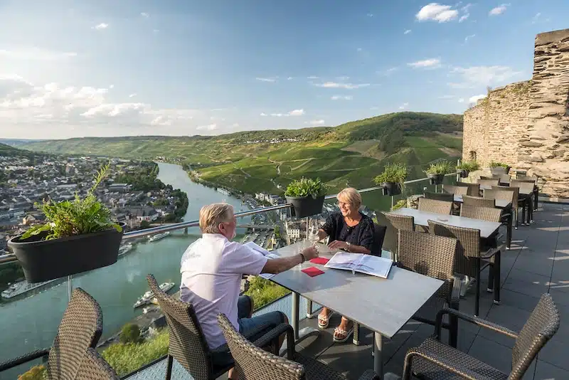 Uit eten met uitzicht op de moezel. man en vrouw aan tafeltje dat uitzicht biedt op de moezel en de heuvels met daaronder de stad.