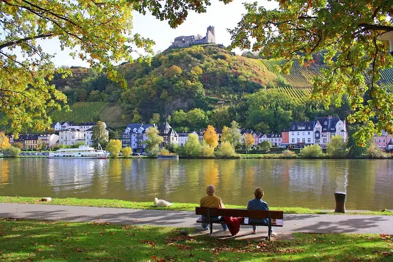 Aan de oever van de Moezel met zicht op Bernkastel-Kues. Mensen zitten op een bankje met uitzicht over de moezel met de stad en de groene heuvel met burcht op de achtergrond.